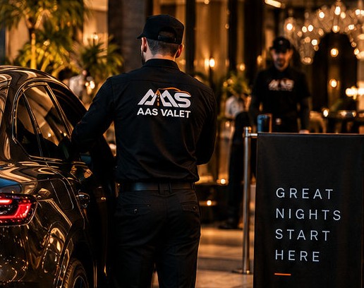 An AAS valet in black uniform opens a guest car at a lit venue entrance at night.